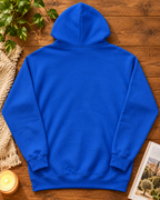 A royal blue hoodie shown from the back, laid flat on a rustic wooden surface with decorative props including a plant, a candle, a woven textile, and an open book.