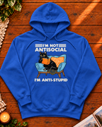 A royal blue pullover hoodie laid flat on a wooden surface, featuring a graphic of a black cat reading an orange book on a light blue armchair with the text 'I'M NOT ANTISOCIAL' above and 'I'M ANTI-STUPID' below.