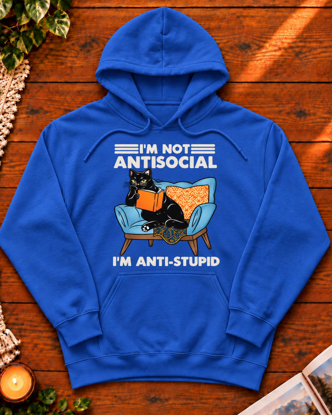 A royal blue pullover hoodie laid flat on a wooden surface, featuring a graphic of a black cat reading an orange book on a light blue armchair with the text 'I'M NOT ANTISOCIAL' above and 'I'M ANTI-STUPID' below.