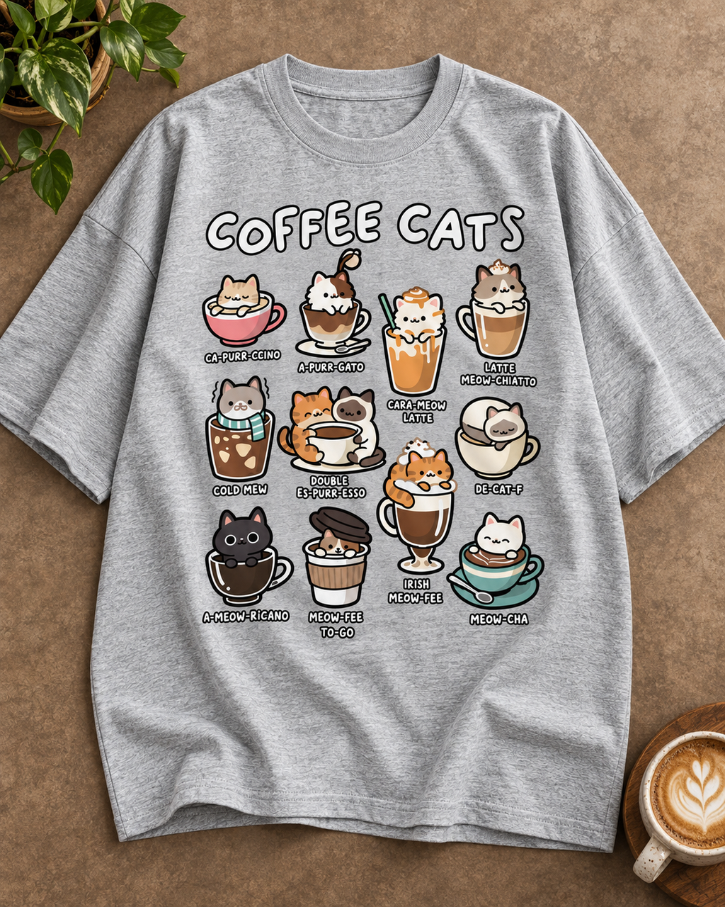A heather gray short-sleeve t-shirt with a 'COFFEE CATS' graphic print, laid flat on a textured brown surface with a plant and a cup of latte in the corners.