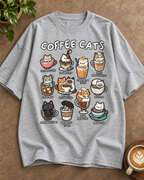 A heather gray short-sleeve t-shirt with a 'COFFEE CATS' graphic print, laid flat on a textured brown surface with a plant and a cup of latte in the corners.